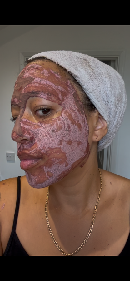 A person wearing a pinkish-purple clay face mask and a white towel wrapped around their hair, wearing a black tank top and a gold chain necklace.