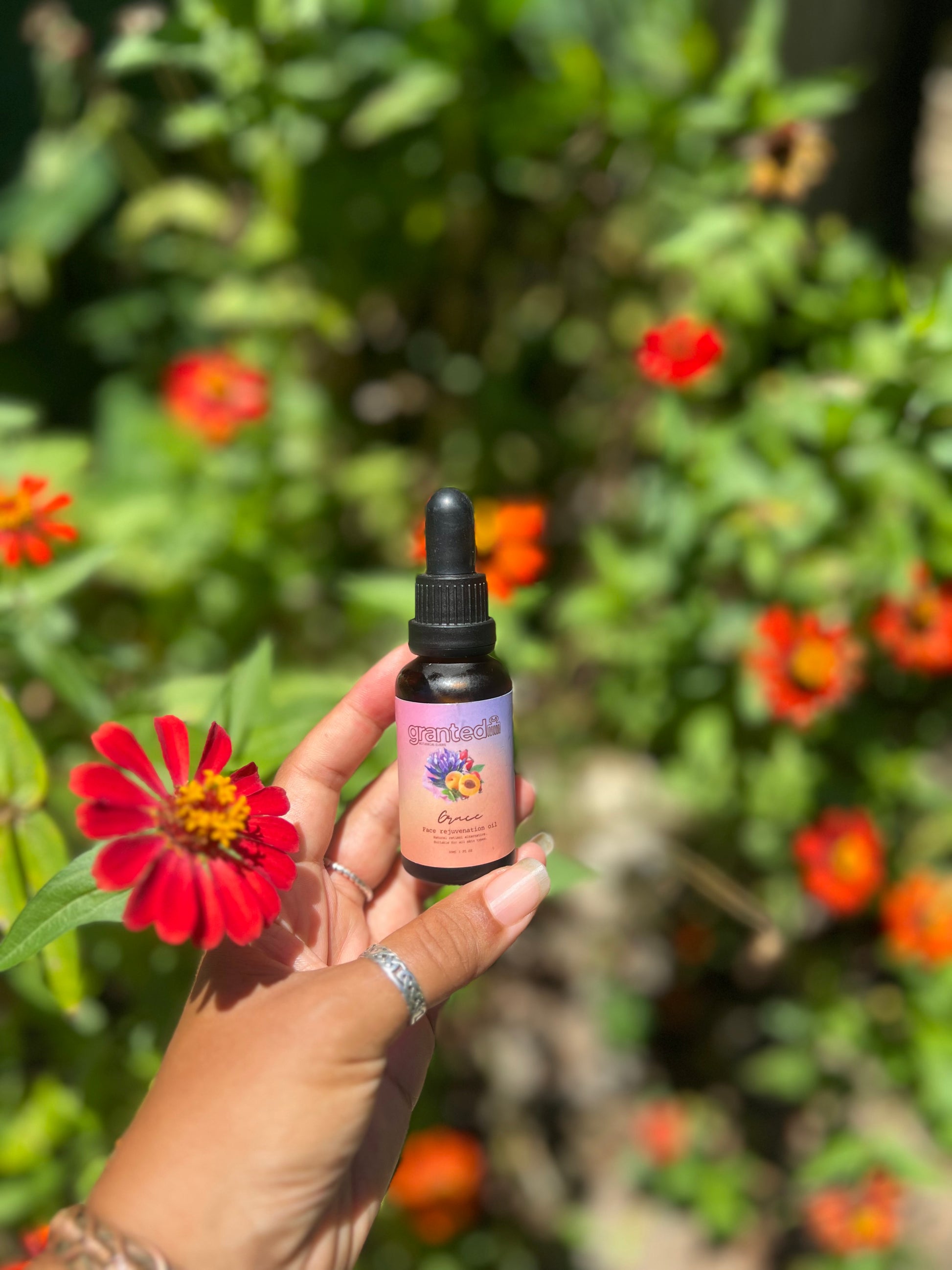 A hand holds a small amber dropper bottle labeled 'Herbs & Flowers' against a blurred garden background with green foliage and red flowers.